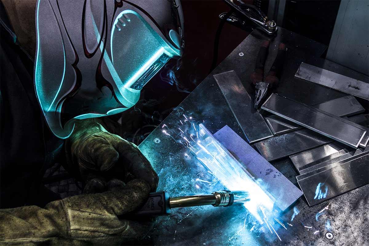 Das Bild zeigt eine Person, die mit einem Schweißhelm und Schutzhandschuhen ausgestattet ist und gerade eine Schweißarbeit durchführt. Der Schweißer verwendet ein WIG-Schweißgerät, das einen hellen Lichtbogen erzeugt. Auf dem Tisch liegen mehrere Metallplatten und Werkzeuge, darunter eine Zange. Die Umgebung ist dunkel, was den blauen Schimmer des Schweißlichts hervorhebt.