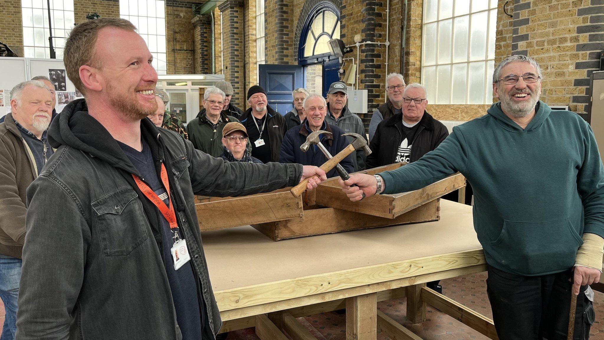 The image shows a group of men gathered in what appears to be a workshop or industrial setting. Two men in the foreground are smiling and holding hammers, crossing them in a gesture of camaraderie or celebration. The man on the left is wearing a black jacket and has a lanyard with an ID badge. The man on the right is wearing a green hoodie and has a bandage on his left wrist. Behind them, several other men are standing, some wearing hats and glasses, all appearing to be in good spirits. The background features brick walls, large windows, and a wooden workbench, suggesting a workshop environment.