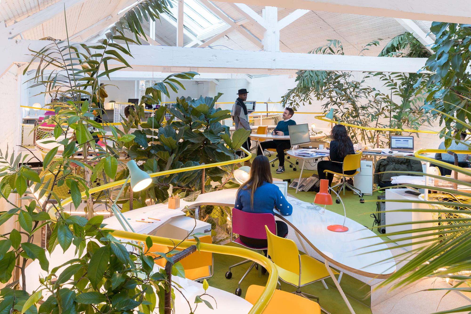 The image depicts a modern, open-plan office space filled with lush green plants, creating a vibrant and natural atmosphere. The workspace features several desks with colorful chairs, including pink and yellow, and is equipped with computers and office supplies. The ceiling is high with white beams, and natural light floods the area through large skylights. Several people are working and interacting, contributing to a collaborative environment. The presence of plants and bright colors suggests a focus on improving mental health and well-being in the workplace.