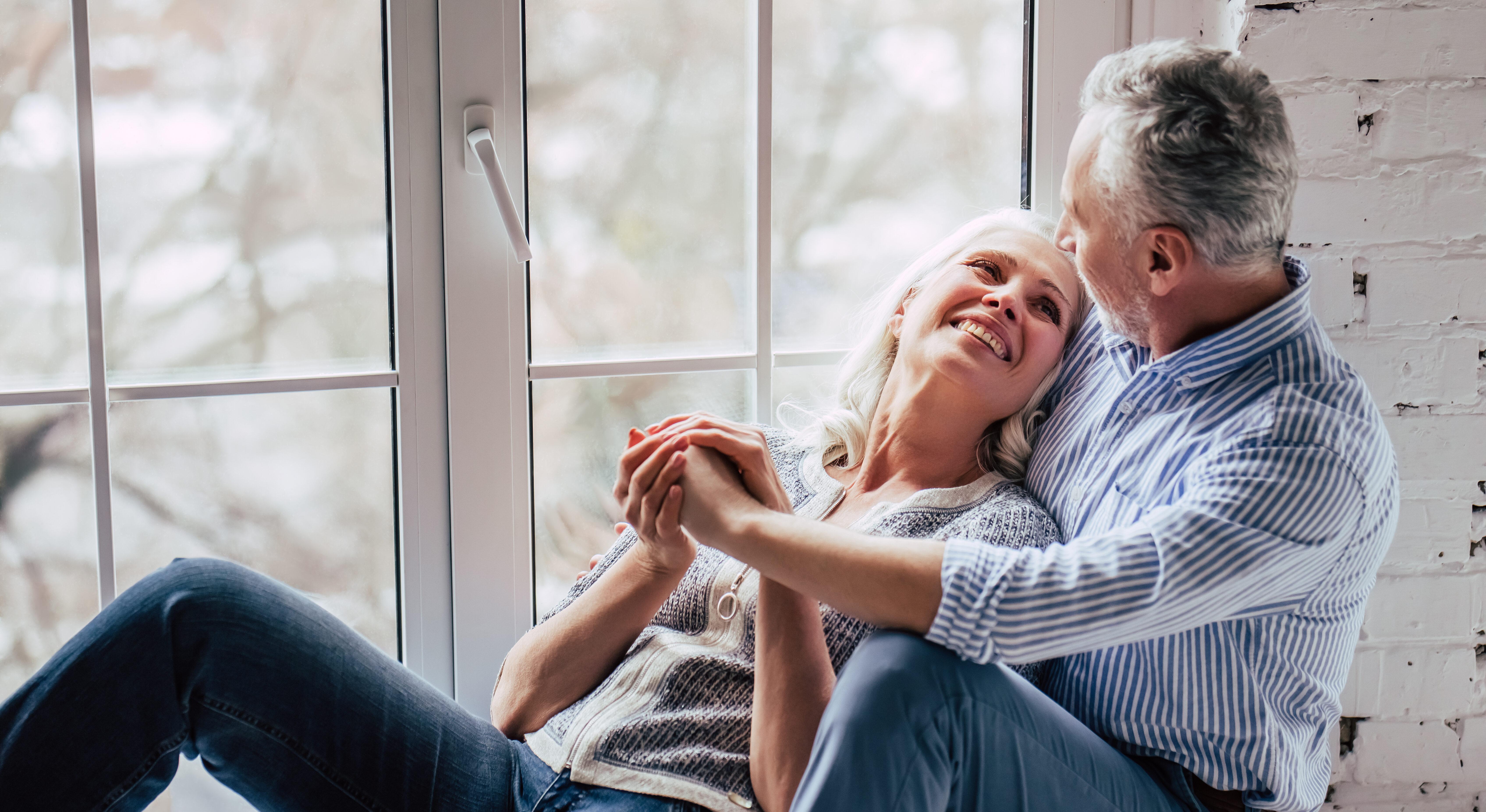 The image depicts a mature couple sitting together by a large window. They appear to be in a relaxed and affectionate pose, with the woman leaning against the man and smiling up at him. The man is looking at her with a gentle expression. The setting is indoors, with a white brick wall partially visible on the right side. The window behind them shows a blurred view of trees, suggesting a serene and peaceful environment. Both individuals are casually dressed, and the overall atmosphere is warm and intimate.
