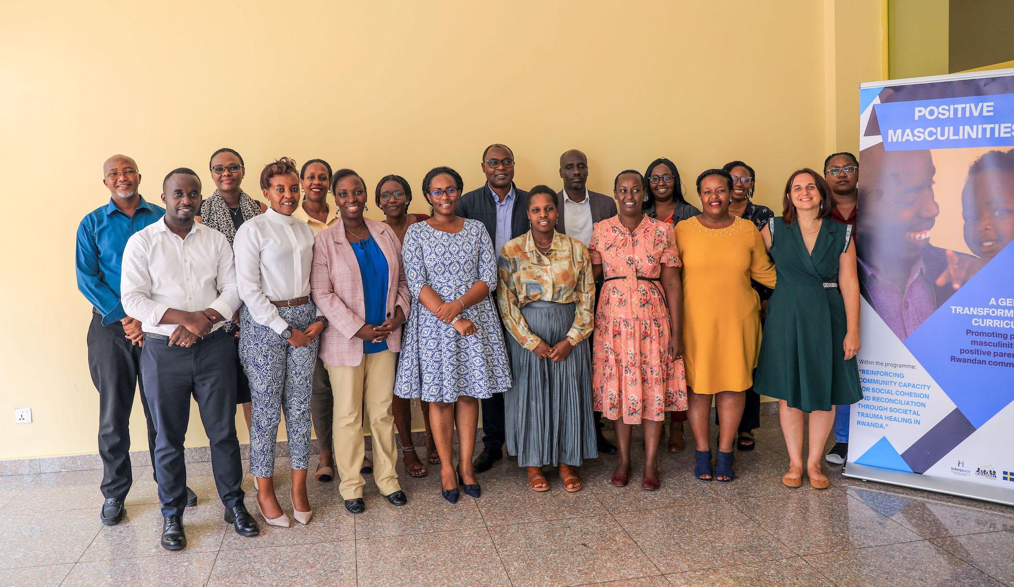 The image shows a group of people standing together in a room with a light-colored wall. There are 18 individuals, both men and women, dressed in formal and semi-formal attire. They are posing for a group photo, smiling at the camera. To the right of the group, there is a vertical banner with the text 'POSITIVE MASCULINITIES' at the top. The banner includes additional text about promoting positive masculinity and parenting within Rwandan communities. The setting appears to be a professional or educational event focused on social themes.