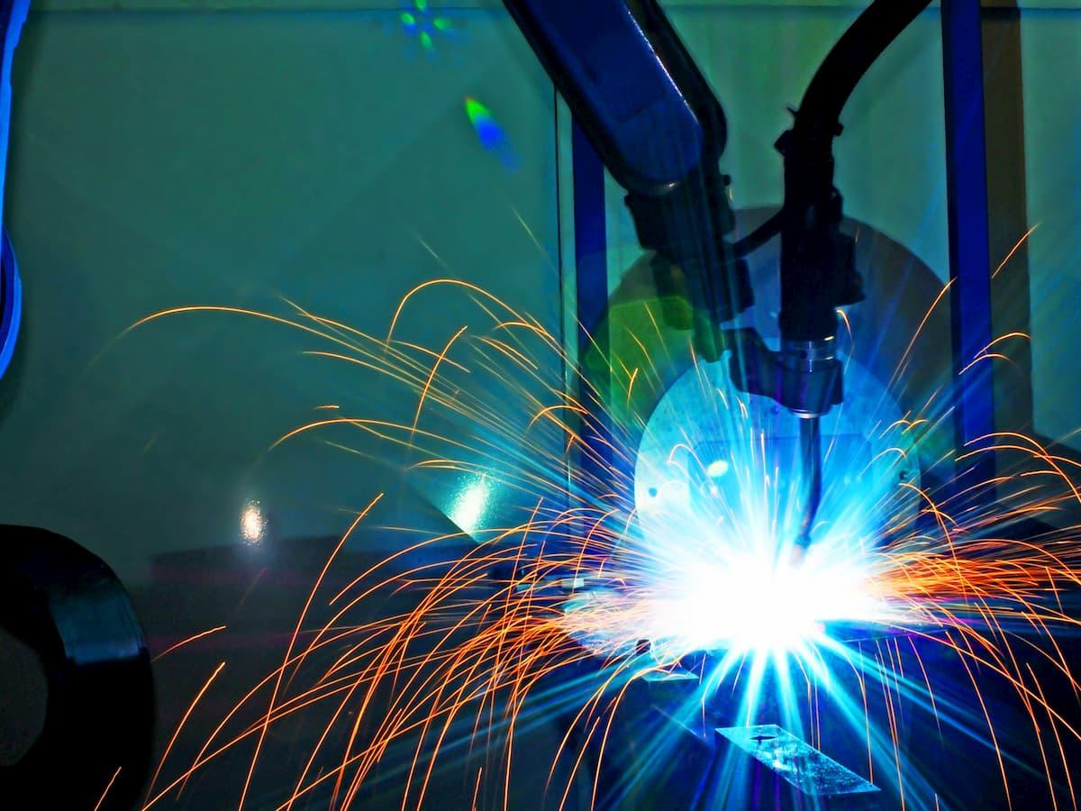 Das Bild zeigt einen industriellen Schweißroboter bei der Arbeit. Der Roboterarm ist in der Mitte des Bildes zu sehen, während er einen hellen Lichtbogen erzeugt, der von blauen und orangefarbenen Funken umgeben ist. Diese Funken fliegen in verschiedene Richtungen, was auf die Intensität des Schweißprozesses hinweist. Der Hintergrund ist unscharf, was den Fokus auf den Schweißvorgang lenkt. Die Szene vermittelt ein Gefühl von Präzision und moderner Technologie in der industriellen Fertigung.