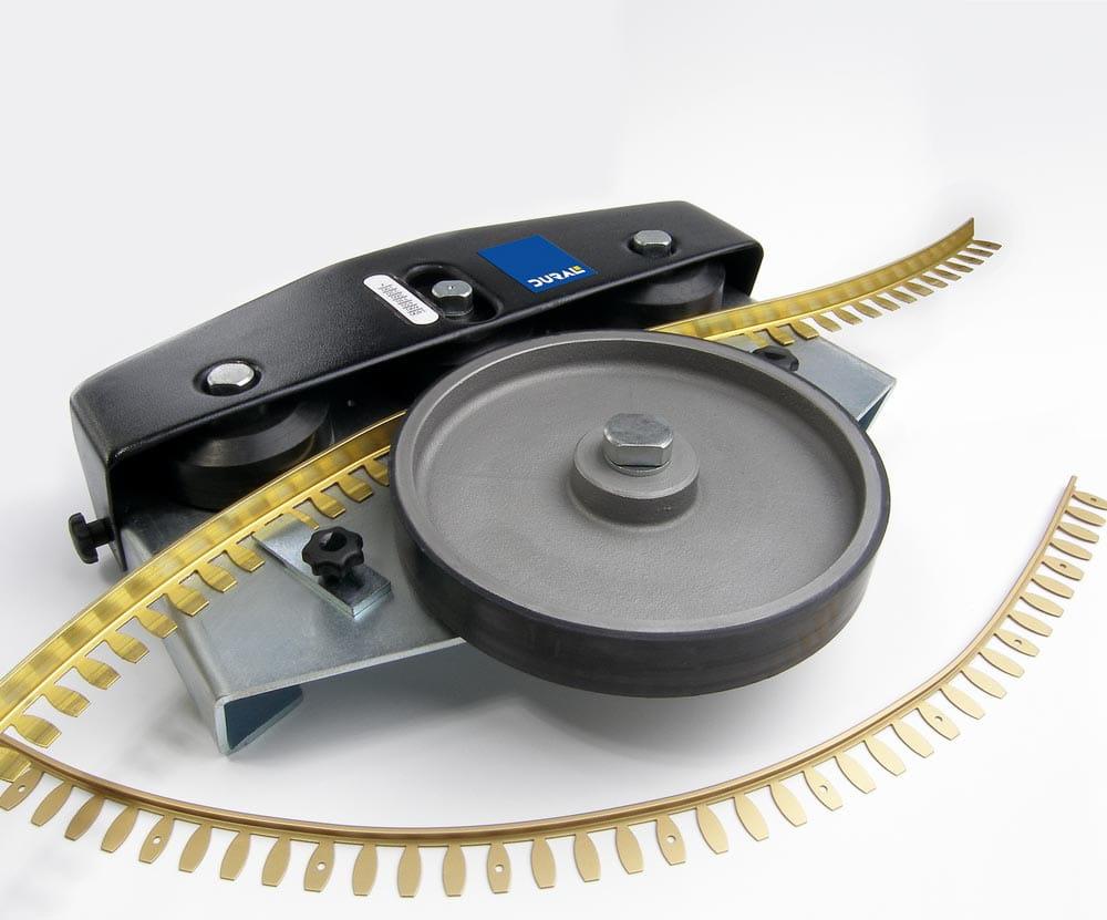 Das Bild zeigt ein Werkzeug, das zum Biegen von flexiblen Fliesenschienen verwendet wird. Im Vordergrund ist eine Metallvorrichtung mit einem großen, runden Rad zu sehen, das auf einer Basis montiert ist. Die Vorrichtung hat eine schwarze Abdeckung mit einem blauen Etikett, auf dem 'DURAL' steht. Um das Werkzeug herum sind gebogene, goldfarbene Fliesenschienen mit regelmäßigen Aussparungen zu sehen, die darauf hindeuten, dass sie flexibel und anpassbar sind. Die gesamte Anordnung ist auf einem weißen Hintergrund dargestellt.