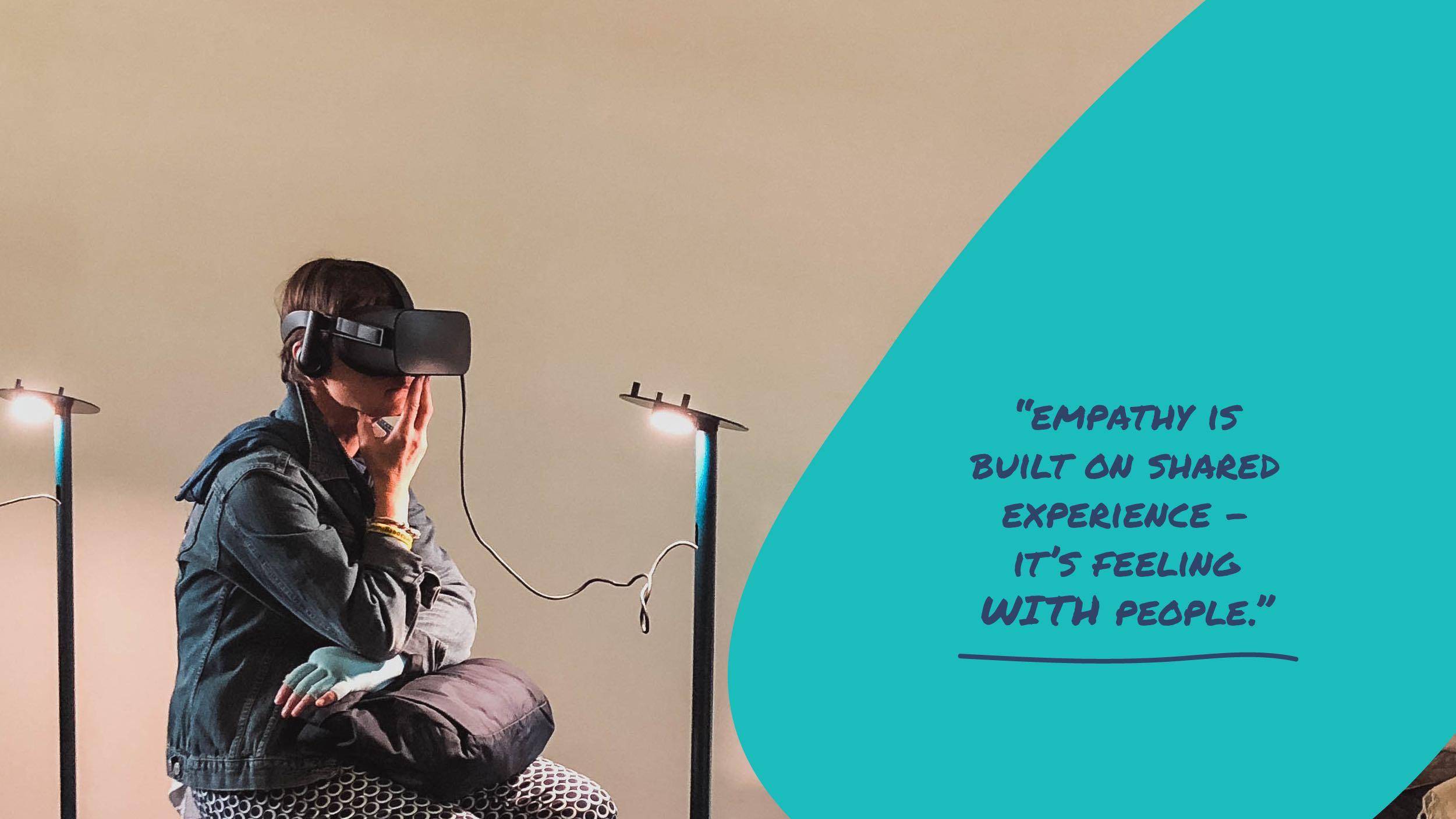 The image shows a person sitting and wearing a virtual reality headset. They are dressed in a denim jacket and patterned pants, with their hand resting on their chin, suggesting a thoughtful pose. Two small lamps are positioned on either side, providing light. On the right side of the image, there is a blue area with text that reads: 'Empathy is built on shared experience - it's feeling with people.' The setting appears to be indoors, with a focus on the concept of using virtual reality to generate empathy.