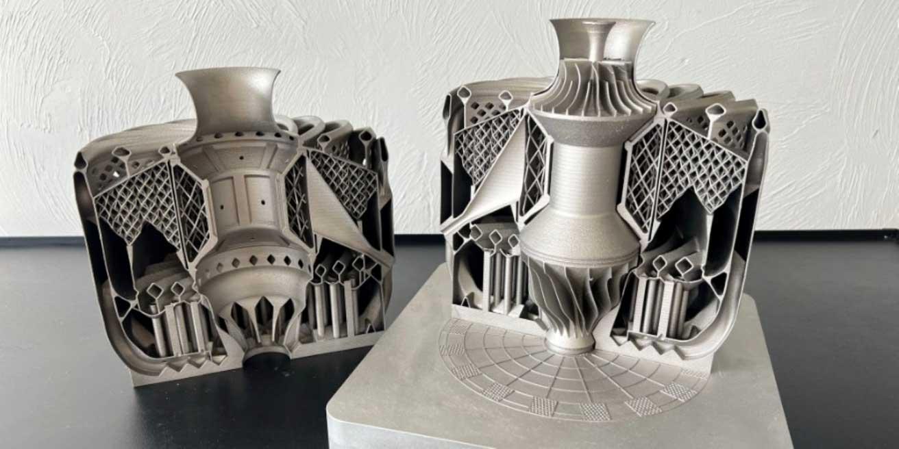 Das Bild zeigt zwei komplexe, metallische 3D-gedruckte Objekte, die auf einem Tisch platziert sind. Beide Objekte haben eine ähnliche Struktur und scheinen technische oder industrielle Komponenten darzustellen. Sie bestehen aus einer Vielzahl von geometrischen Formen, darunter Gitterstrukturen und zylindrische Elemente. Die Oberfläche der Objekte ist glatt und metallisch glänzend. Der Hintergrund ist eine weiße, strukturierte Wand.