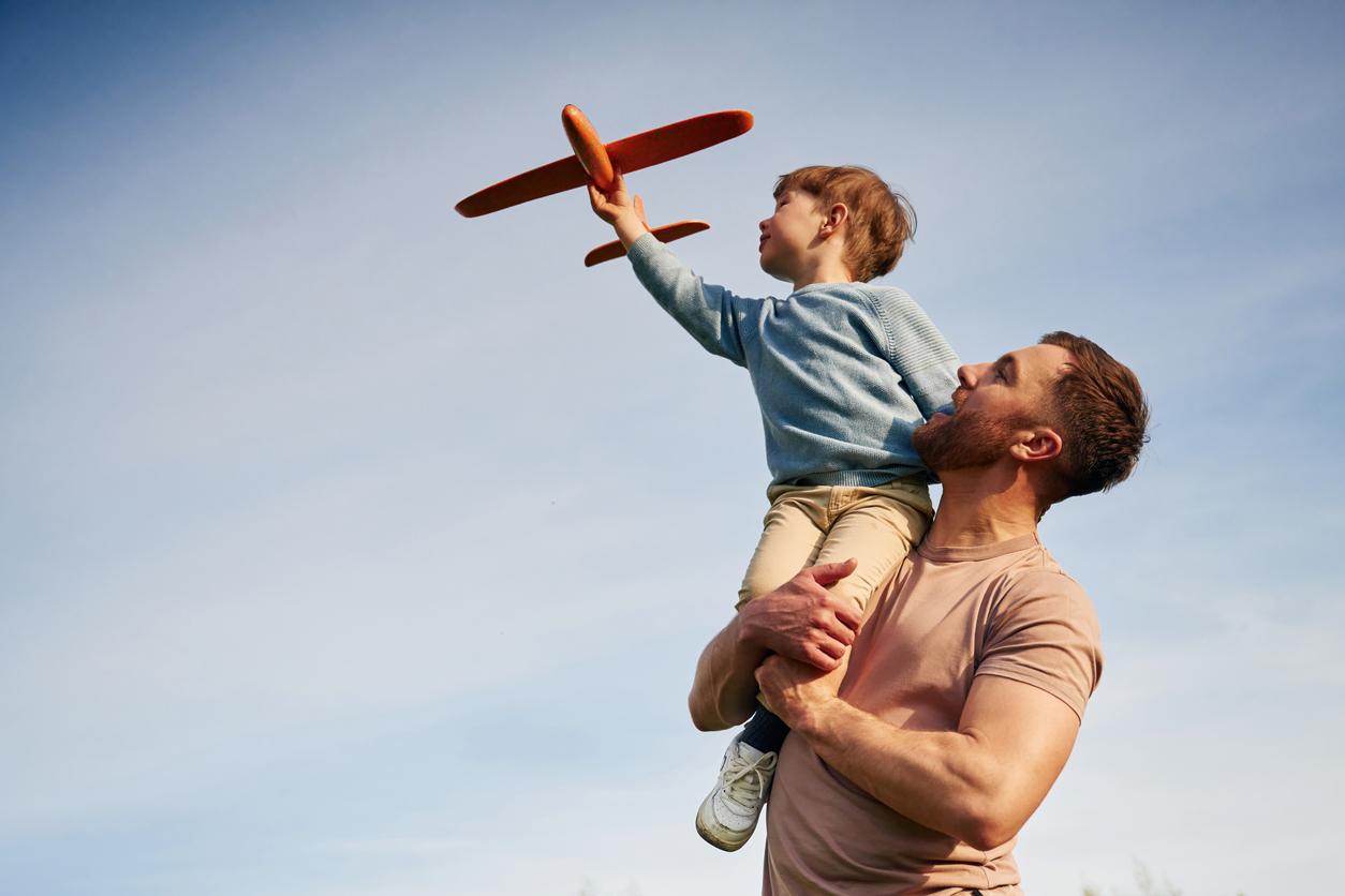 The image depicts a joyful scene of a man holding a young boy on his shoulders. The boy is reaching out with a toy airplane in his hand, seemingly pretending to fly it through the sky. The man is looking up at the boy with a smile, and they are both outdoors under a clear blue sky. The boy is wearing a light blue sweater and beige pants, while the man is dressed in a light pink t-shirt. The atmosphere is playful and warm, suggesting a moment of bonding and imagination.