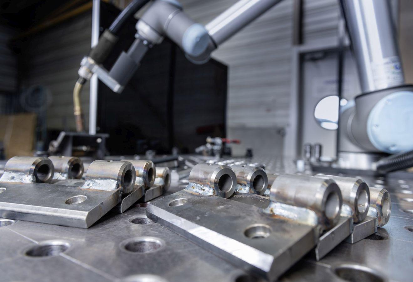 Das Bild zeigt eine industrielle Umgebung mit einem Roboterarm, der über einem Tisch mit Metallteilen arbeitet. Die Metallteile sind auf einer Arbeitsfläche angeordnet, die Löcher aufweist, möglicherweise für Befestigungszwecke. Der Roboterarm scheint für Schweißarbeiten oder ähnliche Aufgaben konzipiert zu sein, da er mit einem Werkzeug ausgestattet ist, das wie eine Schweißdüse aussieht. Im Hintergrund sind unscharfe industrielle Geräte und Strukturen zu erkennen.
