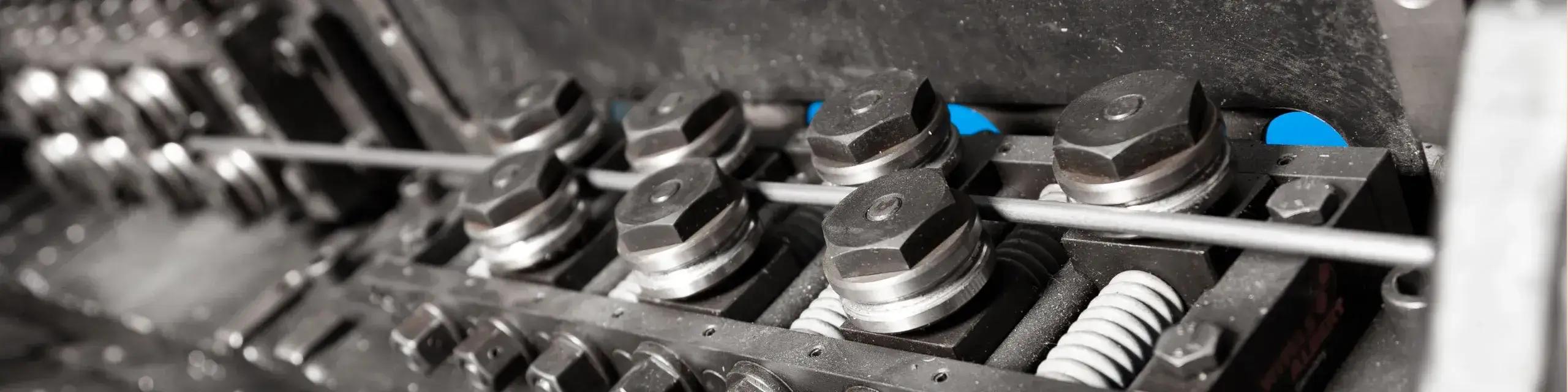 Das Bild zeigt eine Nahaufnahme einer industriellen Maschine, die für das Biegen von Draht verwendet wird. Im Vordergrund sind mehrere große, sechseckige Schrauben oder Muttern zu sehen, die auf einer Metallstange montiert sind. Diese Komponenten scheinen Teil eines Mechanismus zu sein, der den Draht in die gewünschte Form biegt. Die Umgebung ist metallisch und industriell, mit einem Fokus auf Präzision und Technik. Die Farben sind überwiegend in Grautönen gehalten, was auf eine technische und funktionale Umgebung hinweist.