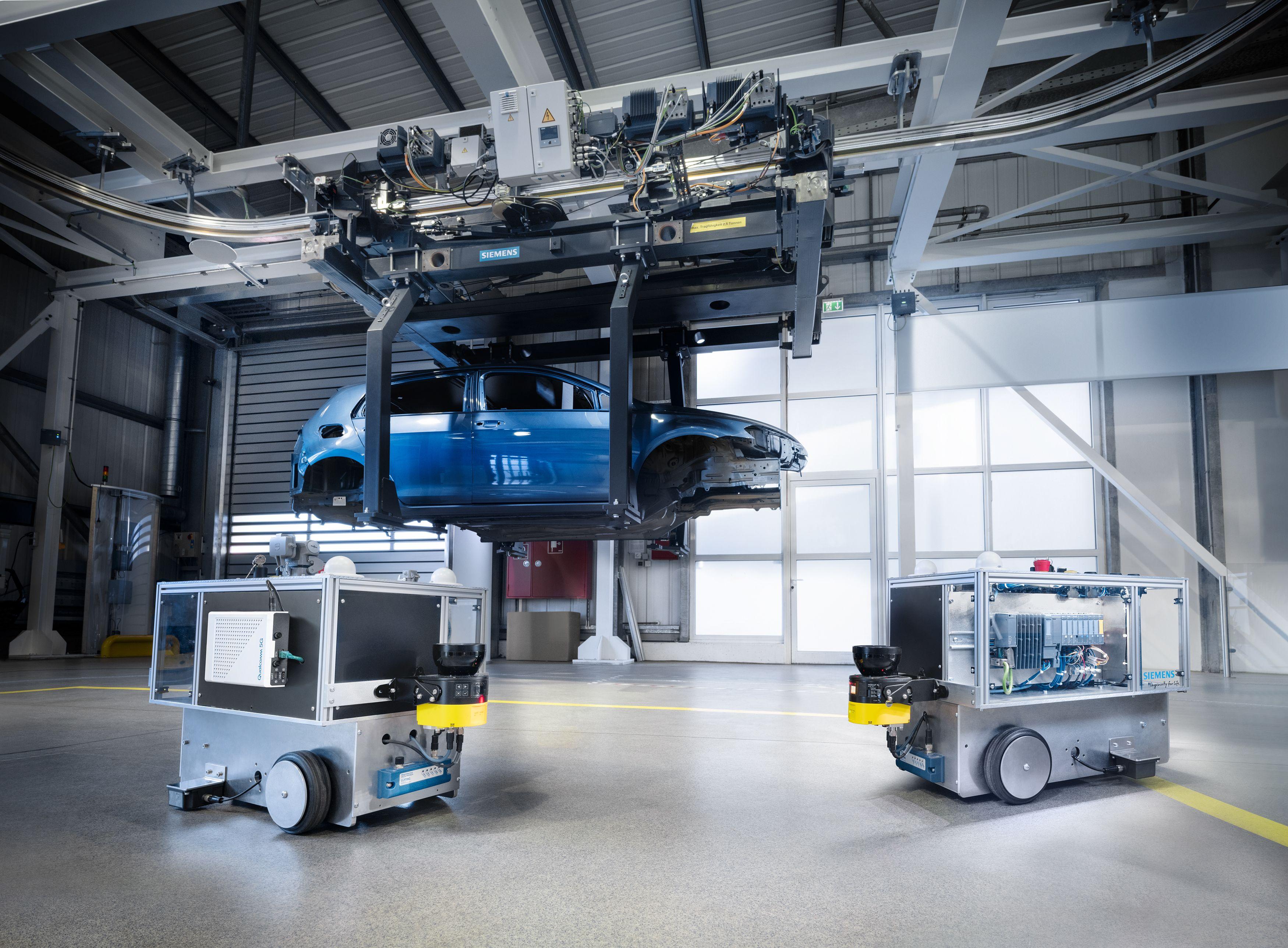Das Bild zeigt eine industrielle Umgebung, in der ein blauer Autokörper an einem automatisierten Fördersystem hängt. Im Vordergrund befinden sich zwei autonome mobile Roboter, die mit elektronischen Komponenten ausgestattet sind. Die Umgebung wirkt modern und technisch fortschrittlich, mit klaren Linien und einer sauberen, gut beleuchteten Werkshalle. Auf dem Fördersystem ist das Logo von Siemens zu erkennen, was auf eine industrielle Automatisierungslösung hinweist.