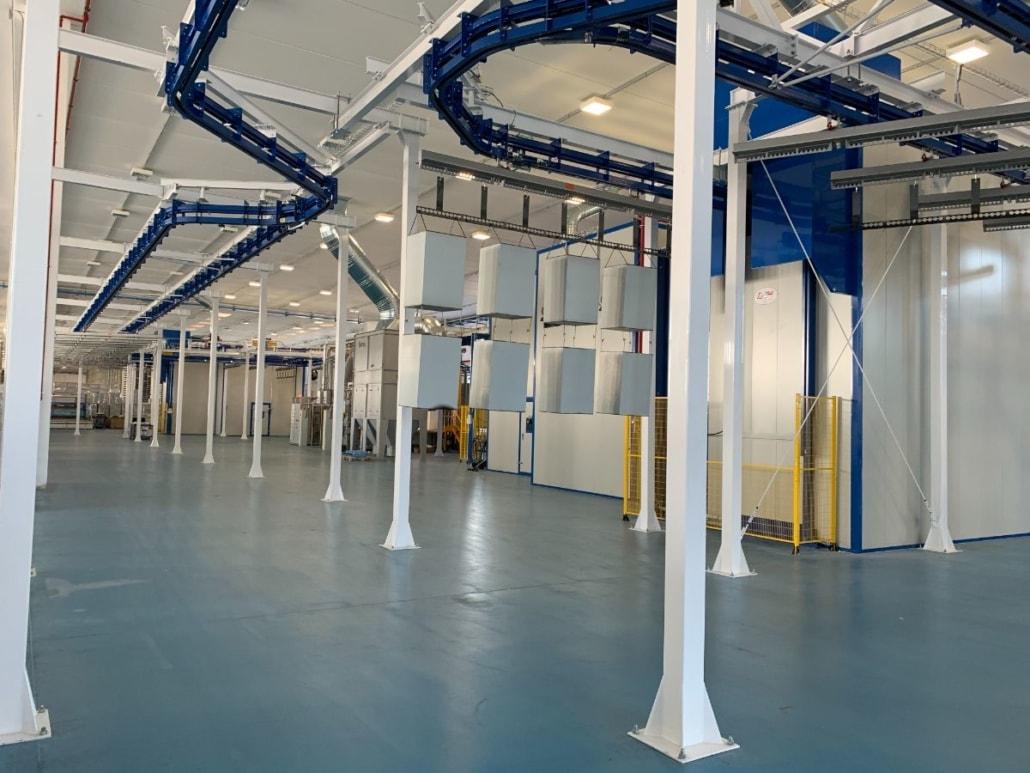 Das Bild zeigt eine große industrielle Halle mit einer automatischen Pulverbeschichtungsanlage. Die Halle ist sauber und gut beleuchtet, mit einem blauen Boden und weißen Wänden. An der Decke sind blaue Förderbänder installiert, die durch den Raum verlaufen. Diese Förderbänder sind Teil der Beschichtungsanlage, die für die Automobilindustrie verwendet wird. Es gibt mehrere weiße Säulen, die die Struktur der Halle unterstützen. Im Hintergrund sind verschiedene Maschinen und Anlagen zu sehen, die Teil des Produktionsprozesses sind.