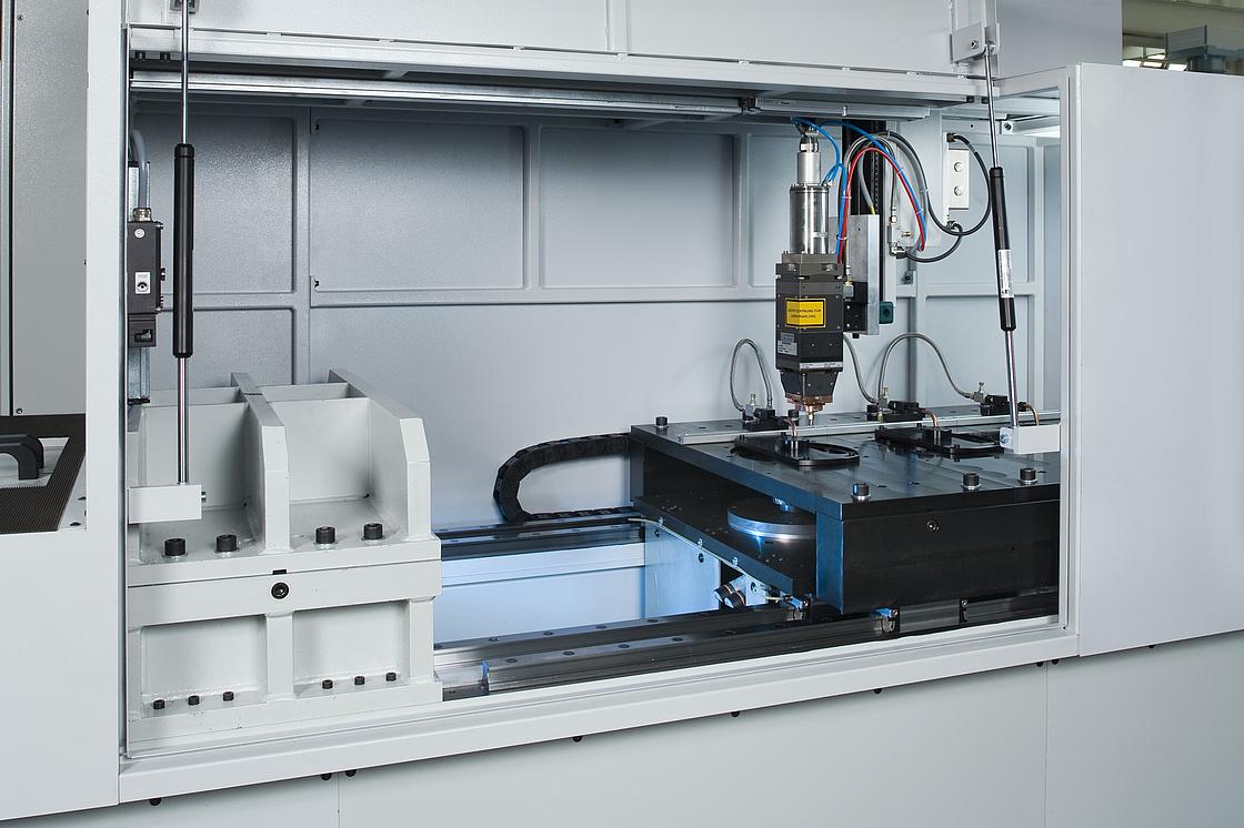 Das Bild zeigt das Innere einer industriellen Lasermaschine. Die Maschine ist in einem hellgrauen Gehäuse untergebracht und enthält verschiedene mechanische und elektronische Komponenten. In der Mitte befindet sich ein Laserkopf, der mit mehreren Kabeln verbunden ist. Der Laserkopf ist auf einer schwarzen Plattform montiert, die sich auf einer Schiene bewegt. Die Umgebung ist sauber und gut organisiert, was auf eine präzise und kontrollierte Arbeitsumgebung hinweist. Die Maschine scheint für die Bearbeitung von Materialien oder Komponenten in der Fertigung eingesetzt zu werden.