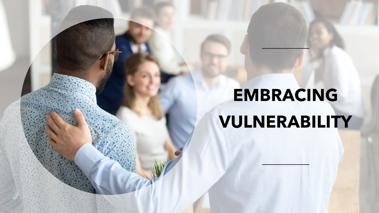 The image shows a group of people in a professional setting, possibly an office or a meeting room. In the foreground, a man with short hair and a light blue shirt has his arm around another man wearing glasses and a patterned shirt, suggesting a gesture of support or camaraderie. In the background, several people are seated and appear to be engaged in conversation, with a focus on a woman smiling. The text 'EMBRACING VULNERABILITY' is prominently displayed on the right side of the image, indicating the theme of the scene.