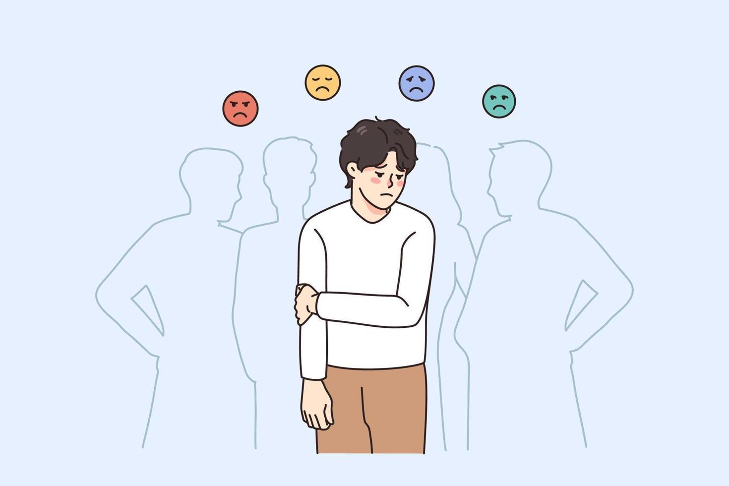 The image depicts a young man standing with a sad expression, holding his arm, suggesting discomfort or loneliness. He is surrounded by the faint outlines of other people, indicating a crowd, but he appears isolated. Above him are four emoticons with different sad expressions, each in a different color: red, yellow, blue, and green. The background is a light blue, enhancing the somber mood of the illustration.