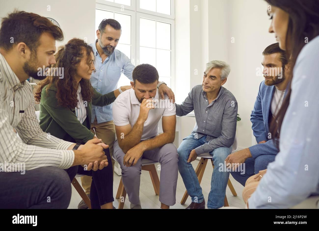 The image depicts a group therapy session where several people are gathered in a supportive manner around a man who appears to be distressed. The man in the center is sitting with his head down, looking contemplative or upset, while others around him offer comfort by placing hands on his shoulders. The setting is a bright room with large windows, and the participants are seated on chairs arranged in a circle. The atmosphere suggests empathy and support, typical of a therapeutic or counseling environment.