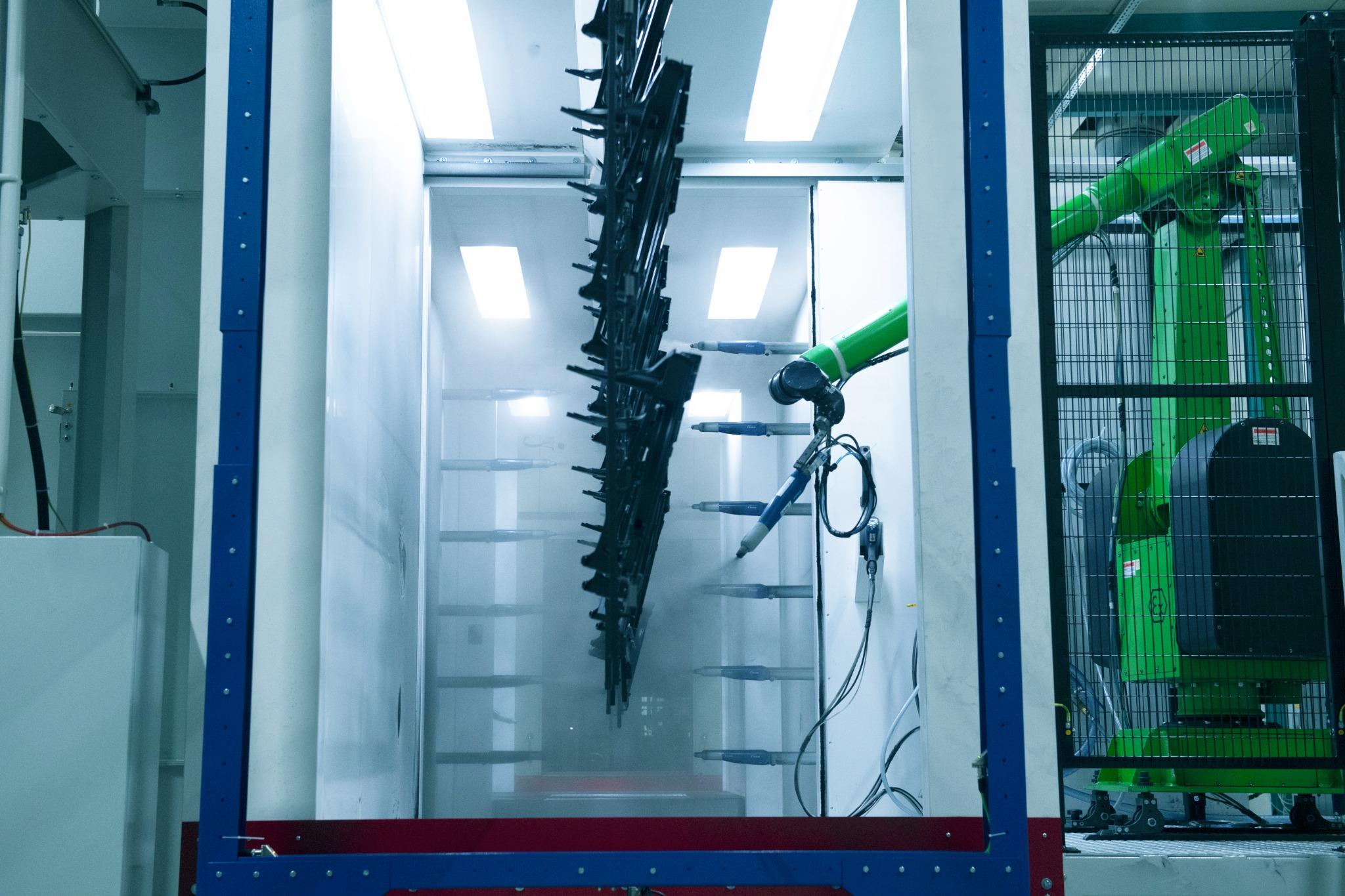 Das Bild zeigt eine industrielle Anlage zur elektrostatischen Pulverbeschichtung. In der Mitte des Bildes befindet sich ein Förderband mit mehreren schwarzen Objekten, die beschichtet werden. Rechts im Bild ist ein grüner Roboterarm zu sehen, der mit einer Sprühvorrichtung ausgestattet ist. Die Umgebung ist hell beleuchtet, und es gibt mehrere Lichtquellen an der Decke. Die Wände der Anlage sind weiß, und es gibt blaue und rote Akzente an der Struktur. Der Roboterarm ist durch ein Gitter geschützt.